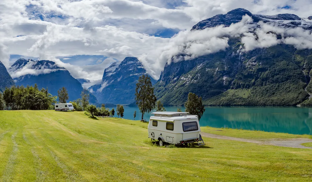 Two RVs parked in a green field next to a beautiful lake and mountains.
