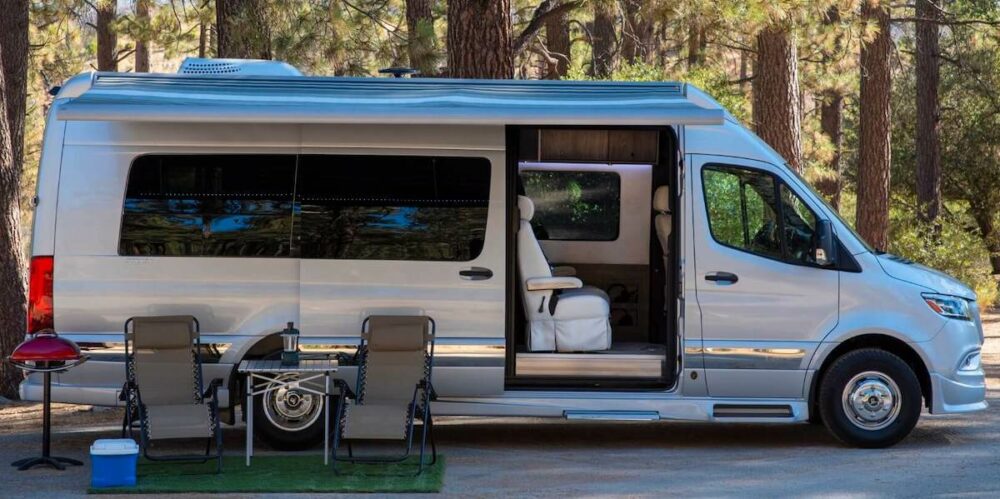 Grech RV Strada Class B RV parked in the woods with the side door open and camping equipment set up outside.